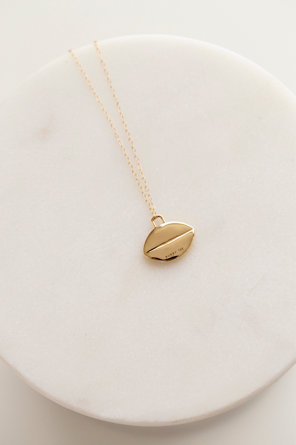 This image shows the back of the flat 14k gold rugby ball pendant. It has no detailing on the back except for a single horizontal line running through the middle of the pendant, and two very small stamps representing the metal quality (14k) and the logo Rugby Love.