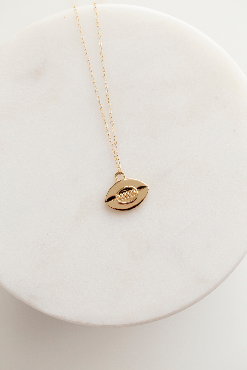 This image shows a 14mm tall by 14.5mm wide flat 14k gold rugby ball pendant with vintage inspired oval stitching detail in the center. Six vertical stitches are set within the oval to mimic the leather stitching of a rugby ball, and there is a horizontal line running through the center of the pendant to symbolize where the two pieces of leather meet on a rugby ball. The bail is a rounded off rectangle set at the top of the flat rugby ball. The pendant is shown on a 1mm gold cable chain.