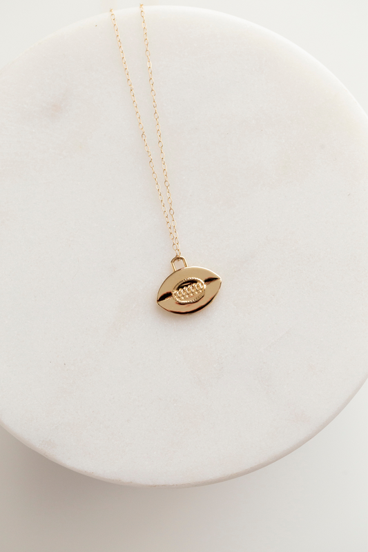 This image shows a 14mm tall by 14.5mm wide flat 14k gold rugby ball pendant with vintage inspired oval stitching detail in the center. Six vertical stitches are set within the oval to mimic the leather stitching of a rugby ball, and there is a horizontal line running through the center of the pendant to symbolize where the two pieces of leather meet on a rugby ball. The bail is a rounded off rectangle set at the top of the flat rugby ball. The pendant is shown on a 1mm gold cable chain.