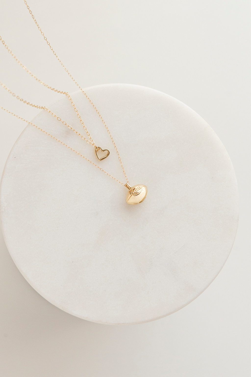 This photo shows two necklaces lying on a round piece of marble. The necklace on the top is a 7mm x 6mm solid gold open heart pendant on a 14k gold cable chain, and the second necklace is the 3d mini puffed rugby ball pendant that features an oval stitching detail with six vertical stitches within the oval. A horizontal line runs through the center of the 3d rugby ball to represent where the two pieces of leather come together on a vintage rugby ball.