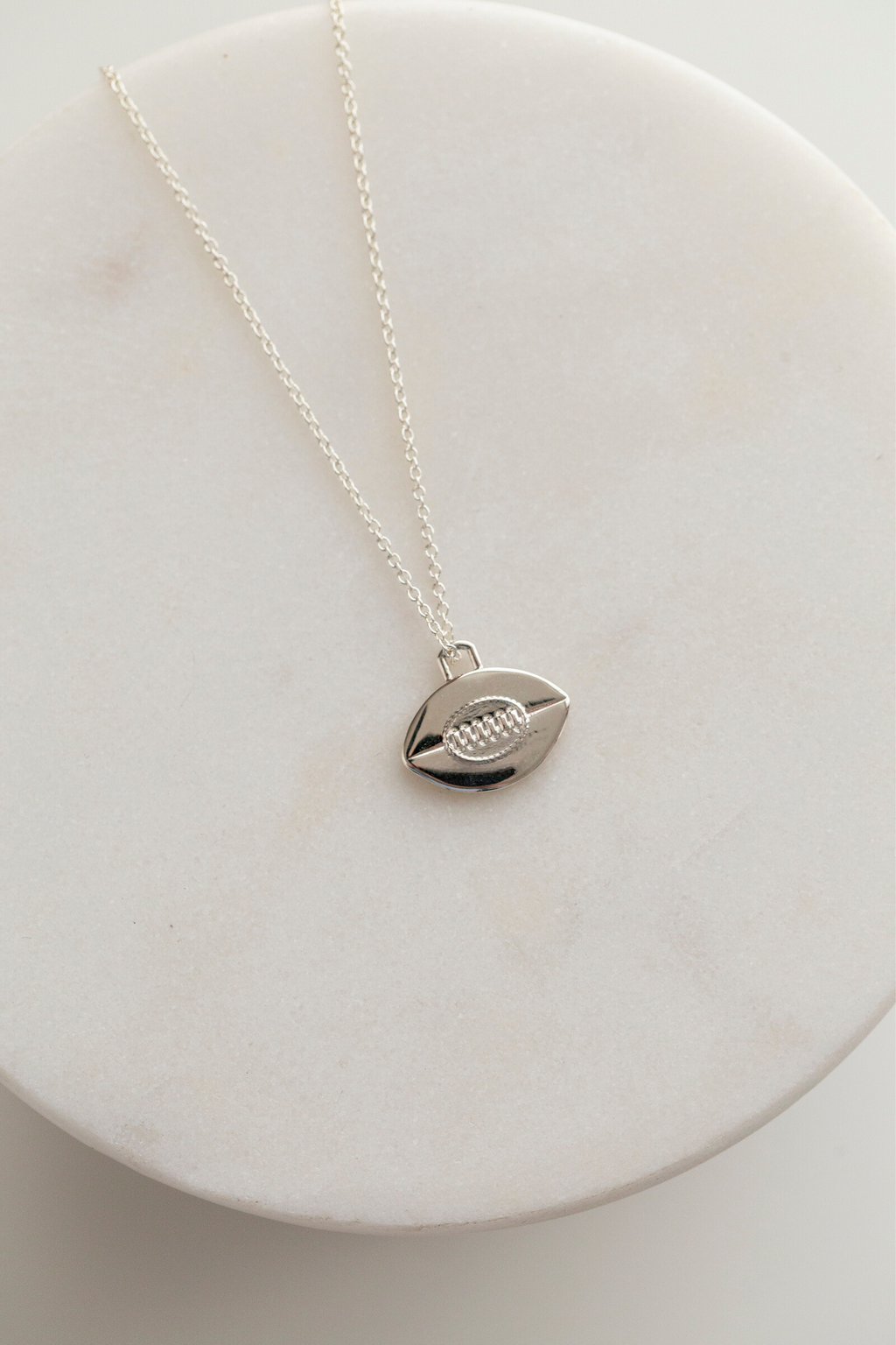 This image shows a 14mm tall by 14.5mm wide flat sterling silver rugby ball pendant with vintage inspired oval stitching detail in the center, six vertical stitches are set within the oval to mimic the leather stitching of a rugby ball, and there is a horizontal line running through the center of the pendant to symbolize where the two pieces of leather meet on a vintage ball. The bail is a rounded off rectangle set at the top of the flat rugby ball. The pendant is shown on a 1mm sterling silver cable chain.