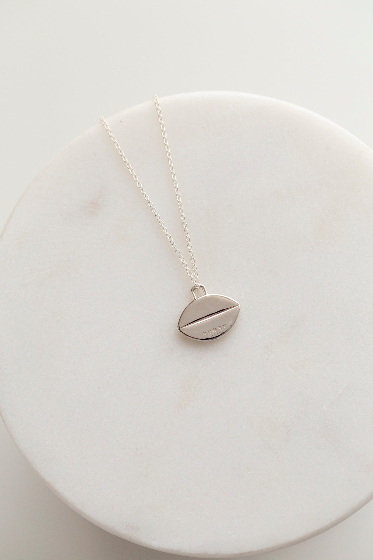 This image shows the back of the flat sterling silver rugby ball pendant. It has no detailing on the back except for a single horizontal line running through the middle of the pendant, and two very small stamps representing the metal quality (sterling) and the logo Rugby Love.